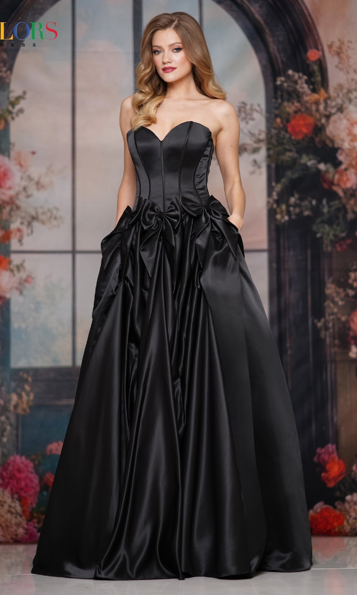 A woman in the Bow-Front Long Prom Ball Gown (Colors Dress 3739) with a fitted bodice and full skirt, poses before flowers and an arched window, embodying timeless Colors Dress elegance.